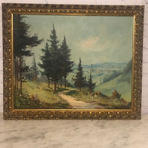 May include: A framed oil painting depicting a landscape scene with a winding path leading through a forest of pine trees towards a distant town. The painting is set against a blue sky with white clouds.