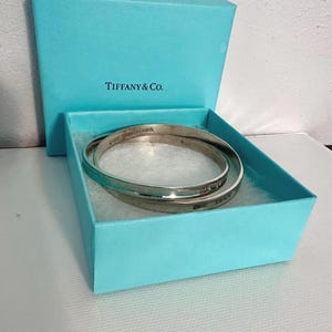 Könnte beinhalten: Zwei silberne Armreifen in einer Tiffany & Co. Box. Die kreisförmigen Armreifen haben eingravierten Text. Die Box ist hellblau und trägt das Tiffany & Co. Logo auf dem Deckel.