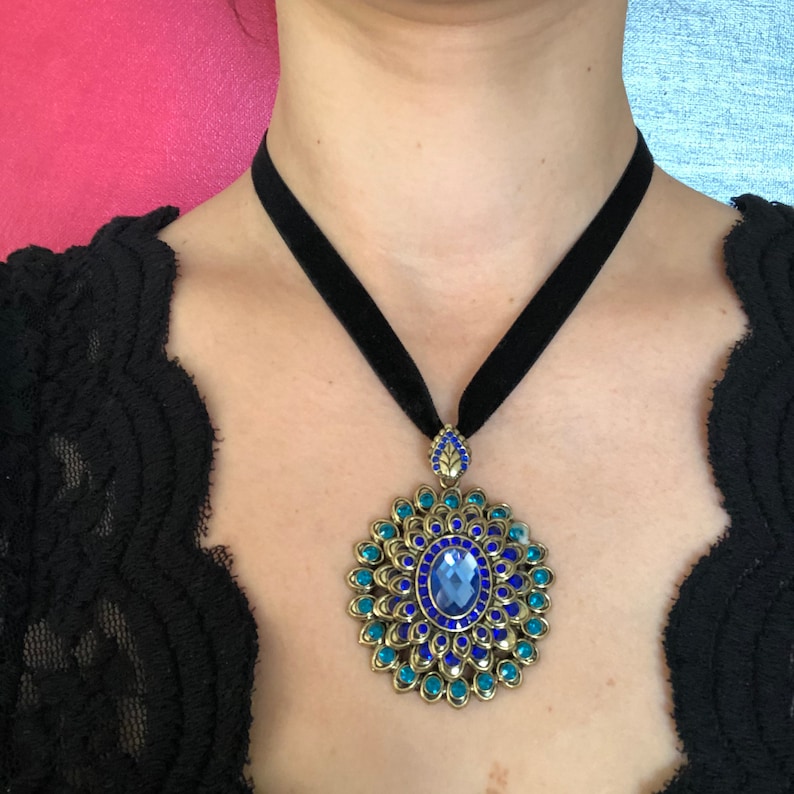 Puede incluir: Un collar gargantilla de terciopelo negro con un colgante dorado que presenta una piedra preciosa azul y turquesa rodeada de un dise&ntilde;o floral decorativo.
