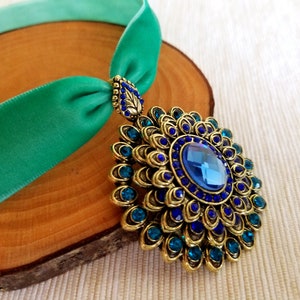 Puede incluir: Un collar gargantilla de terciopelo azul verdoso con un gran colgante de oro y azul adornado con piedras preciosas. El colgante presenta una gran piedra preciosa azul rodeada de piedras preciosas azules m&aacute;s peque&ntilde;as y detalles dorados.
