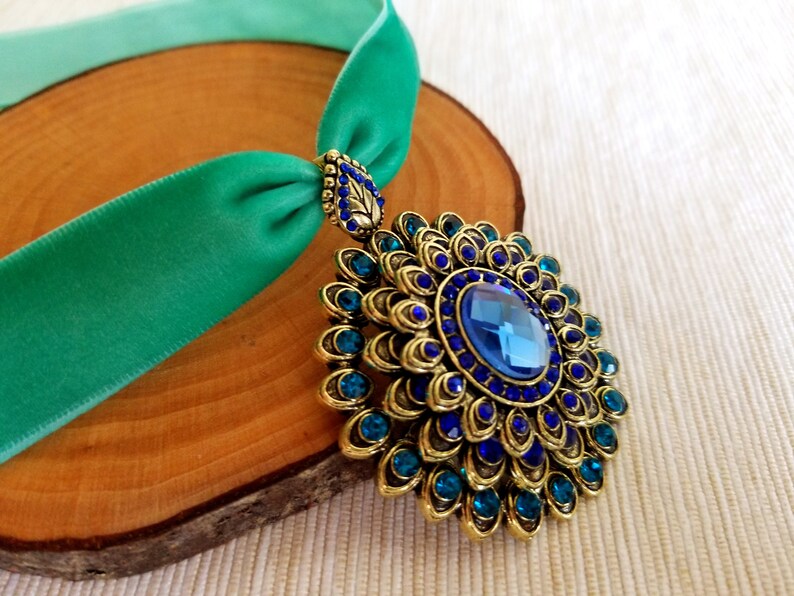 Puede incluir: Un collar gargantilla de terciopelo azul verdoso con un gran colgante de oro y azul adornado con piedras preciosas. El colgante presenta una gran piedra preciosa azul rodeada de piedras preciosas azules m&aacute;s peque&ntilde;as y detalles dorados.
