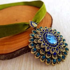 Puede incluir: Un collar gargantilla de terciopelo verde con un gran colgante dorado que presenta una piedra preciosa azul rodeada de piedras preciosas azules y verde azulado m&aacute;s peque&ntilde;as.