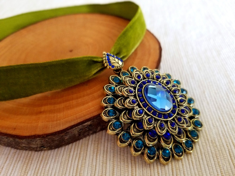 Puede incluir: Un collar gargantilla de terciopelo verde con un gran colgante dorado que presenta una piedra preciosa azul rodeada de piedras preciosas azules y verde azulado m&aacute;s peque&ntilde;as.