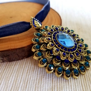 Puede incluir: Un collar con un colgante de oro adornado con una piedra preciosa azul rodeada de peque&ntilde;as piedras azules y turquesa. El colgante tiene la forma de una flor con muchos p&eacute;talos. El collar est&aacute; sobre una cinta de terciopelo azul.