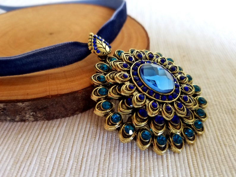 Puede incluir: Un collar con un colgante de oro adornado con una piedra preciosa azul rodeada de peque&ntilde;as piedras azules y turquesa. El colgante tiene la forma de una flor con muchos p&eacute;talos. El collar est&aacute; sobre una cinta de terciopelo azul.