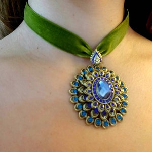 Puede incluir: Un collar gargantilla de terciopelo verde con un gran colgante adornado. El colgante est&aacute; hecho de metal dorado y presenta una gran piedra preciosa azul rodeada de piedras preciosas azules m&aacute;s peque&ntilde;as.