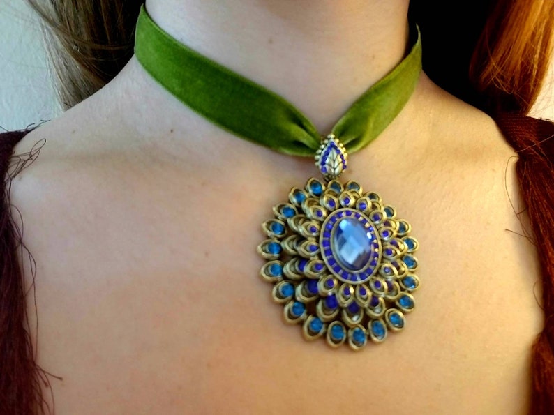 Puede incluir: Un collar gargantilla de terciopelo verde con un gran colgante adornado. El colgante est&aacute; hecho de metal dorado y presenta una gran piedra preciosa azul rodeada de piedras preciosas azules m&aacute;s peque&ntilde;as.