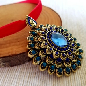 Puede incluir: Un collar con un colgante dorado con una piedra preciosa azul rodeada de piedras preciosas azules y verdes m&aacute;s peque&ntilde;as. El colgante est&aacute; unido a una cinta de terciopelo rojo.