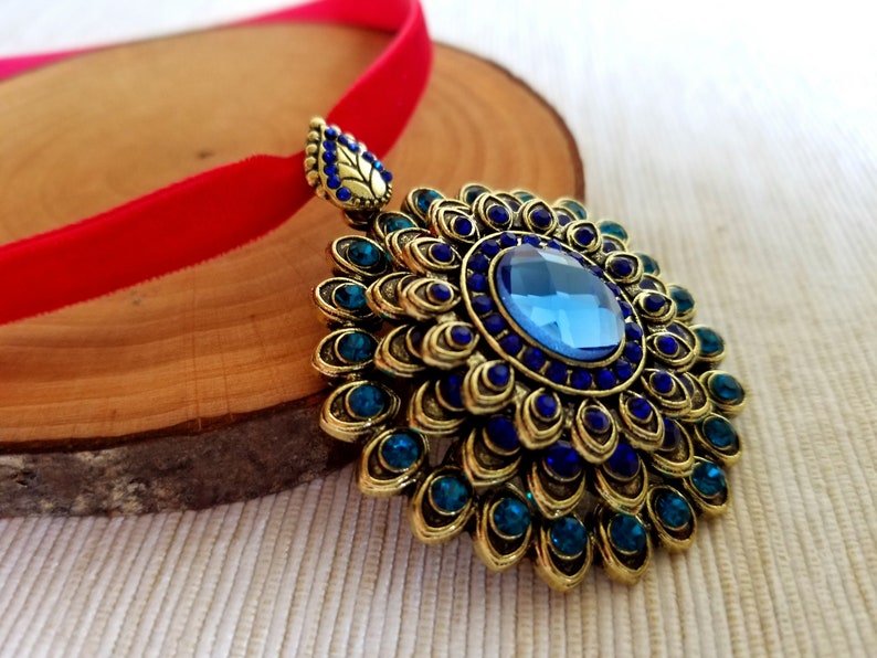 Puede incluir: Un collar con un colgante dorado con una piedra preciosa azul rodeada de piedras preciosas azules y verdes m&aacute;s peque&ntilde;as. El colgante est&aacute; unido a una cinta de terciopelo rojo.