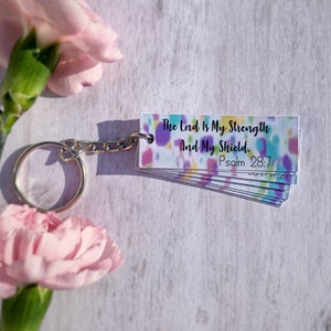 May include: A keychain featuring a stack of small rectangular cards with a colourful, speckled design. The cards display the text "The Lord Is My Strength And My Shield. Psalm 28:7". A silver chain and ring are attached. Pink flowers are in the background.