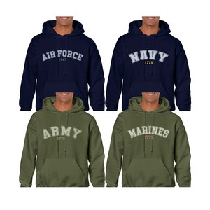 K&ouml;nnte beinhalten: Vier Kapuzenpullover in verschiedenen Farben mit den Namen der Zweige des US-Milit&auml;rs auf der Vorderseite. Die Pullover sind marineblau, olivgr&uuml;n und dunkelgr&uuml;n. Der Text auf den Pullovern lautet "Air Force 1947", "Navy 1775", "Army 1775" und "Marines 1775".