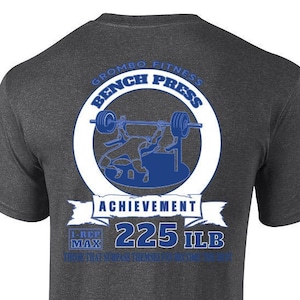 Peut inclure: Un t-shirt gris foncé avec un graphique circulaire blanc au dos. Le graphique représente une silhouette bleue d'une personne soulevant des poids sur un banc. Le texte "Grombo Fitness Bench Press Achievement 225 ILB 1 REP MAX" est imprimé en blanc et bleu sur le graphique.