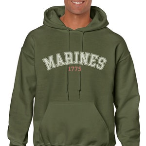 K&ouml;nnte beinhalten: Ein gr&uuml;ner Kapuzenpullover mit dem Wort "MARINES" in Wei&szlig; mit rotem Rand und dem Jahr "1775" darunter.
