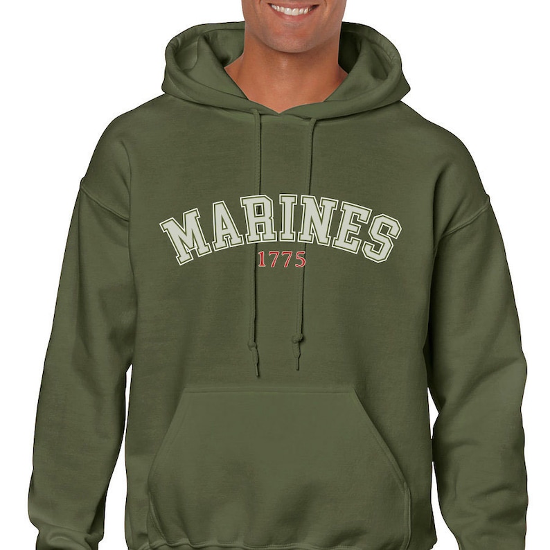 K&ouml;nnte beinhalten: Ein gr&uuml;ner Kapuzenpullover mit dem Wort "MARINES" in Wei&szlig; mit rotem Rand und dem Jahr "1775" darunter.
