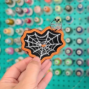 May include: A black and orange embroidered patch featuring a white spider web design. The patch has a scalloped orange border and the text "Wretched Mischief" with a bat logo. The patch is held in a hand.
