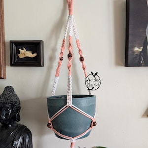 May include: A pink and white macrame plant hanger with wooden beads. The hanger is holding a teal pot. The text "Wretched Mischief" is visible on the hanger.