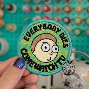 May include: Embroidered patch with a cartoon character's face, surrounded by text that reads "EVERYBODY DIES COME WATCH TV." The patch has a green border and a light green background. The character has tan skin and brown hair.