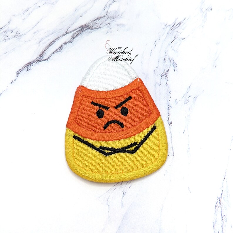 May include: Embroidered candy corn patch with an angry face. The patch features a white top, orange centre, and yellow base, with black embroidered details for the face and outline. The text "Wretched Mischief" is in the upper right corner.