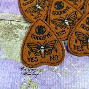 May include: Brown felt planchette patches with black embroidered designs. Each patch features a butterfly, eye, and the words "Goodbye", "Yes", and "No". The patches are in a triangular shape, with a rounded top. The brand name "Wretched Mischief" is also visible.