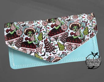 May include: A light blue clutch wallet with a flap closure. The flap features a repeating pattern of a zombie-like woman holding a candy cane, with the text "Undead Debbie". The design includes candy, trees, and bats.