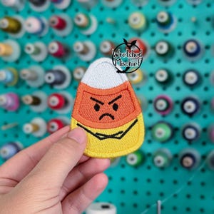May include: Embroidered candy corn patch with an angry face. The patch features white, orange, and yellow colors with black detailing. The patch is held in front of a background of sewing spools. The text "Wretched Mischief" is above the patch.