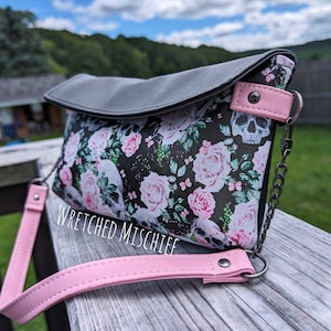 May include: A black and white floral print clutch bag with a gray flap and pink strap. The bag has a silver chain strap and a skull design. The bag has the text "Wretched Mischief" printed on the front.
