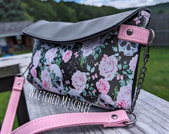 May include: A black and white floral print clutch bag with a gray flap and pink strap. The bag has a silver chain strap and a skull design. The bag has the text "Wretched Mischief" printed on the front.