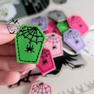 May include: Green felt coffin with a black spider web and spider embroidered on it. The coffin is surrounded by other felt coffins in various colors.