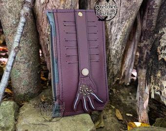May include: A burgundy leather wallet with a zipper closure and a decorative hand design. The wallet has multiple card slots and a zippered pocket. The text "Wretched Mischief" is printed on the wallet.
