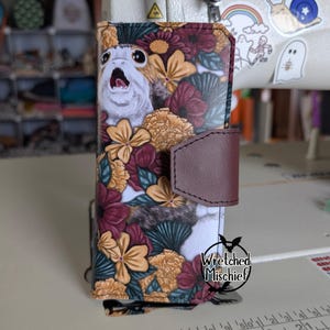 May include: A phone case featuring a floral design with a Porg character. The case has a brown leather clasp and the text "Wretched Mischief" at the bottom. The case is covered in a pattern of flowers in shades of yellow, burgundy, and green.