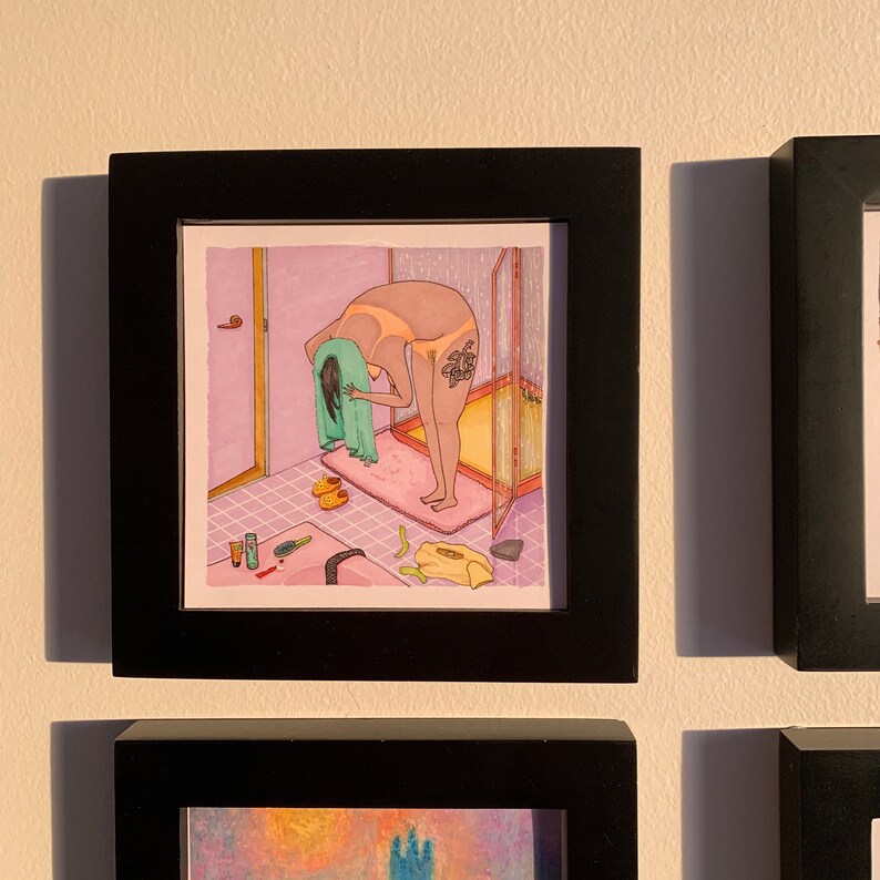 Puede incluir: Una impresi&oacute;n art&iacute;stica enmarcada que representa a una persona en un ba&ntilde;o, sec&aacute;ndose el pelo con una toalla verde. La ilustraci&oacute;n presenta una combinaci&oacute;n de colores rosa y amarillo, con detalles como una ducha, zapatillas y art&iacute;culos de tocador.
