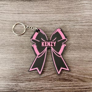 Può includere: Un portachiavi a forma di fiocco nero e rosa con il nome "KENZY" in lettere bianche. Il portachiavi ha un anello e una catena argentati. Il design del fiocco ha contorni rosa e un interno nero. Il portachiavi è su una superficie di legno.