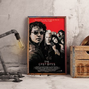 May include: A framed movie poster for "The Lost Boys" with a red background and images of the cast. The poster is displayed next to a metal pipe lamp with a vintage bulb and a wooden crate.