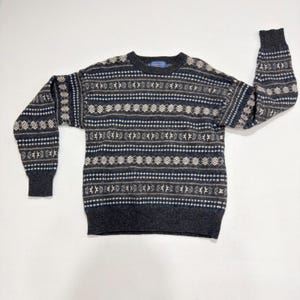May include: A long-sleeve crew neck sweater featuring a dark charcoal gray base with a repeating horizontal pattern of geometric shapes in shades of navy blue, light gray, and cream. The garment has ribbed cuffs, a ribbed hem, and a blue neck label.