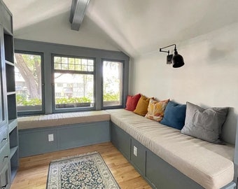 May include: A window seat with a built-in bench and cushions. The bench is covered in a light beige fabric and has a row of colorful throw pillows. The window seat is in a room with a light gray wall and a wooden floor.