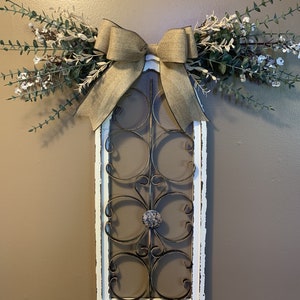 May include: A decorative wall hanging featuring a distressed white frame with a wrought iron design. A burlap bow and faux greenery with white flowers adorn the top. The frame is approximately 76 cm tall.