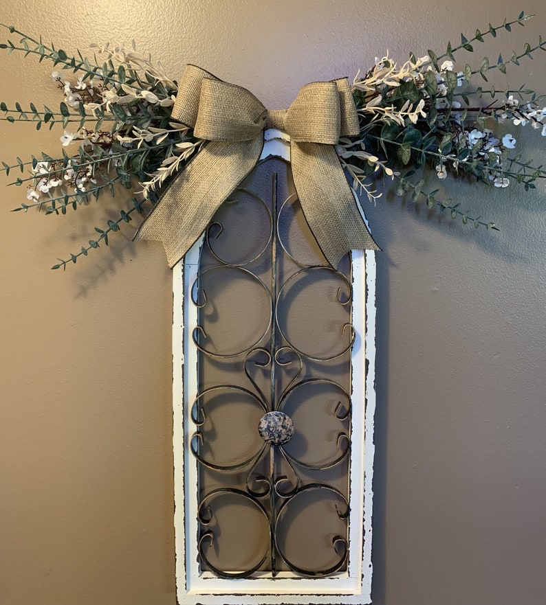 May include: A decorative wall hanging featuring a distressed white frame with a wrought iron design. A burlap bow and faux greenery with white flowers adorn the top. The frame is approximately 76 cm tall.