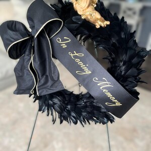 以下が含まれることがあります： 黒い羽根のリース、黒いリボンと金のトリム付き。金色の文字で「In Loving Memory」と書かれた黒いリボン。小さな金のケルビムの置物がリースの上にあります。リースは金属製のスタンドに飾られています。