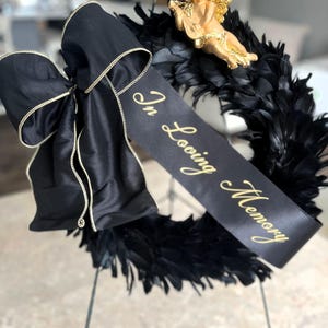 以下が含まれることがあります： 黒い羽根のリース、黒いサテンのリボン、金色の文字で「In Loving Memory」と書かれたバナー。小さな金のケルビム像がリースの上に置かれています。リースは金属製のスタンドに飾られています。