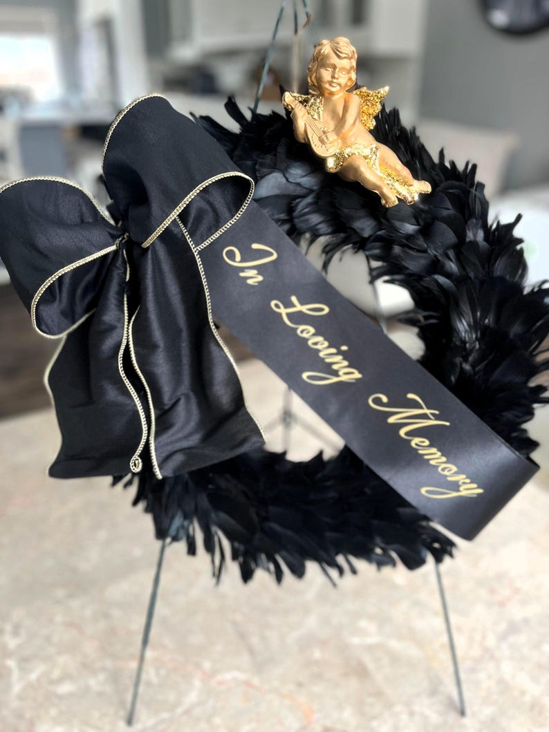 以下が含まれることがあります： 黒い羽根のリース、黒いサテンのリボン、金色の文字で「In Loving Memory」と書かれたバナー。小さな金のケルビム像がリースの上に置かれています。リースは金属製のスタンドに飾られています。