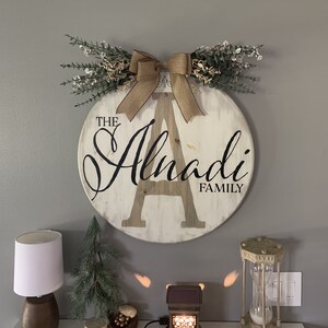 May include: A circular wooden family sign, featuring the text "The Alnadi Family" in black script, with a large central letter "A". It's adorned with a burlap bow and foliage. The sign is mounted on a grey wall.