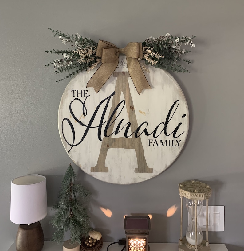 May include: A circular wooden family sign, featuring the text "The Alnadi Family" in black script, with a large central letter "A". It's adorned with a burlap bow and foliage. The sign is mounted on a grey wall.