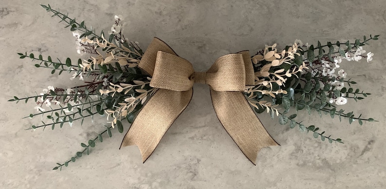 May include: A decorative floral arrangement with a large burlap bow with a dark brown trim. Green and white leafy sprigs extend outwards from the bow. Suitable for home decor, this arrangement would make a lovely addition.