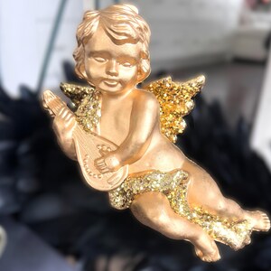以下が含まれることがあります： 金色のケルビムの置物で、リュートを演奏しています。ケルビムはキラキラ光る翼とアクセントを持っています。この置物は、休日や特別な機会のための装飾品かもしれません。