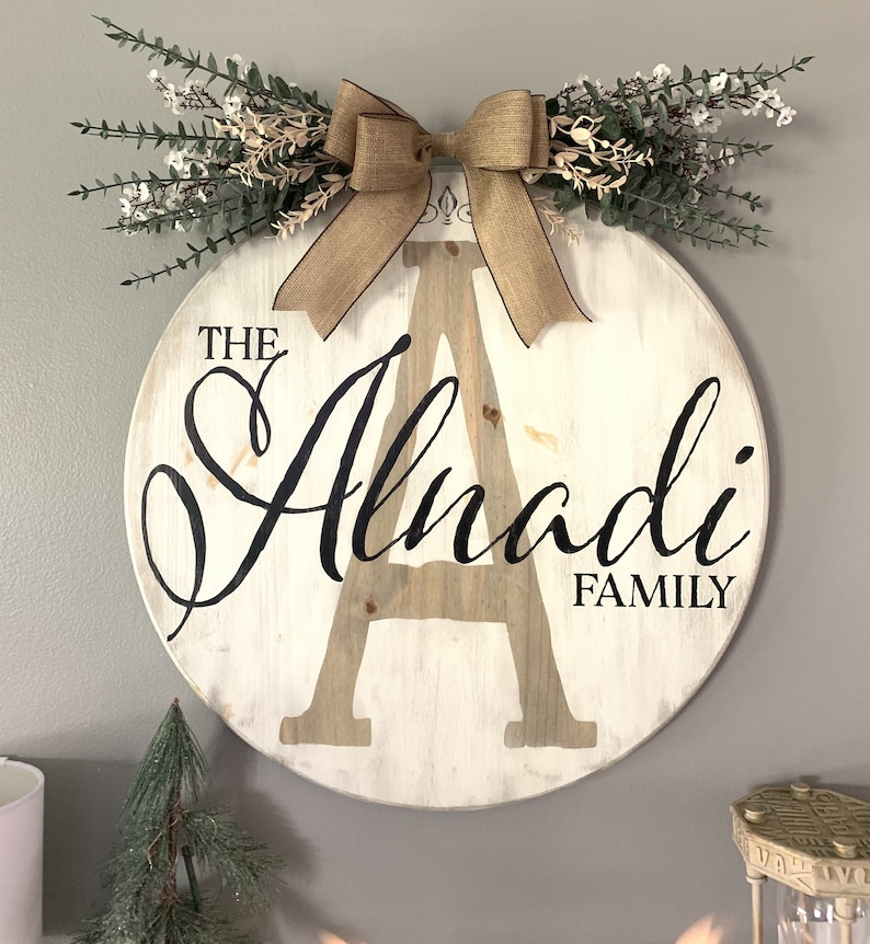 May include: A circular wooden family sign featuring the name "Alnadi" in black script and a large letter "A". It is adorned with a burlap bow and greenery. The words "THE" and "FAMILY" are also visible on the sign.