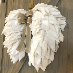 Pu&ograve; includere: Ali d'angelo in tessuto bianco con un fiocco in juta marrone, appese a una porta di legno.