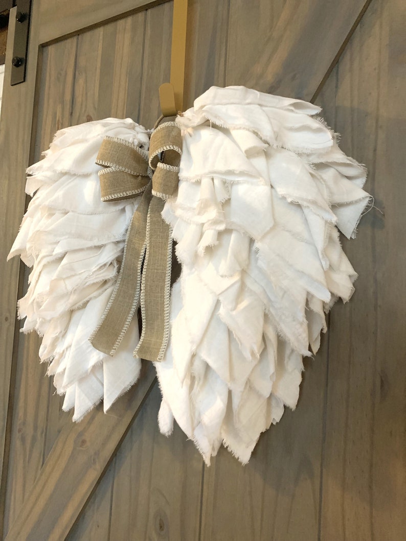 Pu&ograve; includere: Ali d'angelo in tessuto bianco con un fiocco in juta marrone, appese a una porta di legno.