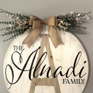 May include: A circular, white wooden family sign featuring the name "Alnadi" in black script. It's decorated with a burlap bow and greenery. The word "FAMILY" is printed below. This sign is designed for home decoration, with a rustic aesthetic.