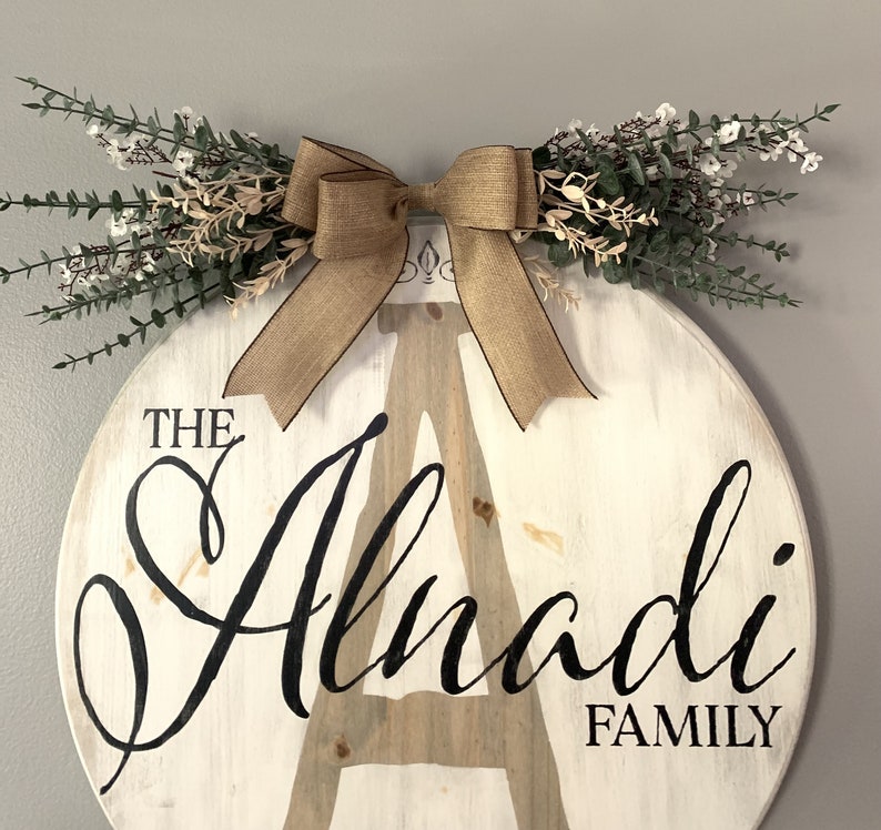 May include: A circular, white wooden family sign featuring the name "Alnadi" in black script. It's decorated with a burlap bow and greenery. The word "FAMILY" is printed below. This sign is designed for home decoration, with a rustic aesthetic.