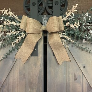 May include: A rustic burlap bow with faux greenery and white flowers, attached to wooden barn doors with metal hardware. The design has a farmhouse aesthetic. The doors are painted in a light grey colour, with a natural wood grain.